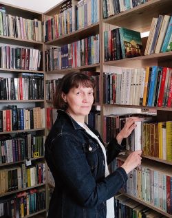 Beata Łyczewska, kobieta w średnim wieku w kręconych włosach do ramion stoi przed regałem pełnym książek