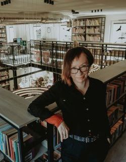 Kamilla Marchewska, brunetka w okularach opiera się o poręcz bibliotecznej antresoli, wokół regały z książkami