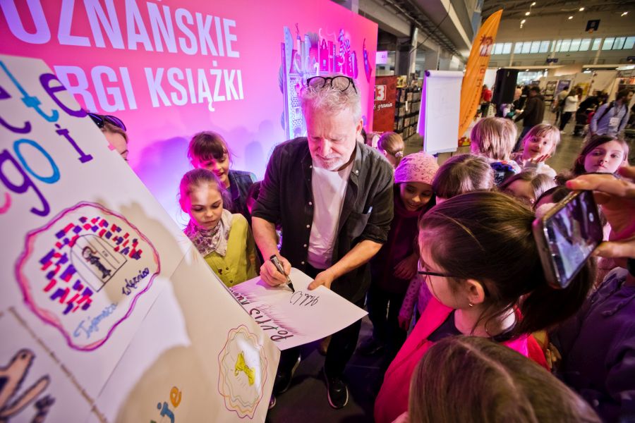fot. Fotobueno Pisarz Martin Widmark podpisuje plakat, otoczony przez grupę dzieci na Targach Książki w Poznaniu. W tle pomarańczowe stoisko z napisem 'Poznańskie Targi Książki