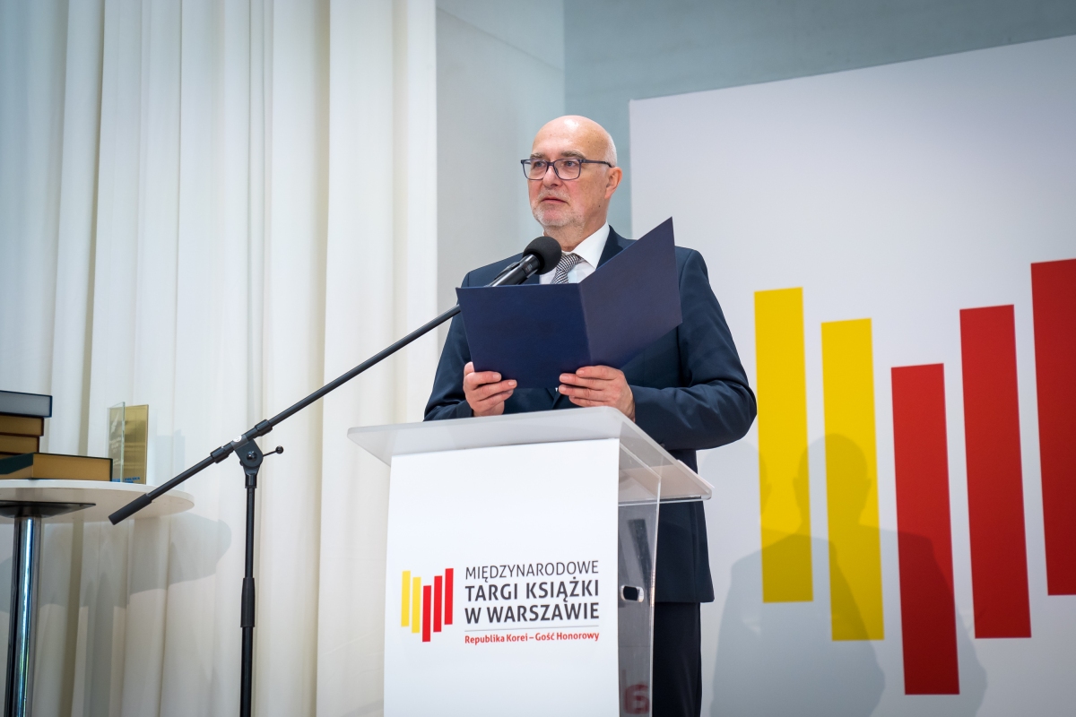Ubrany w garnitur mężczyzna stoi przed mikrofonem. W tle logo targów